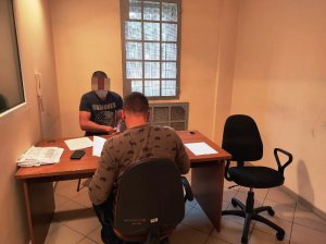 Na zdjęciu znajduje się biurko, przy którym siedzi zatrzymany mężczyzna oraz policjant ubrany po cywilnemu.