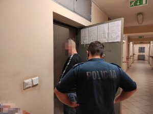 Na zdjęciu widoczny jest zatrzymany 42-latek wraz z umundurowanym policjantem. Zdjęcie zostało wykonana w wejściu do policyjnej celi.