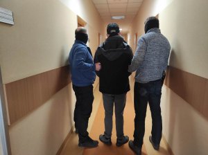 Zdjęcie przestawia trzech mężczyzn, dwaj to policjanci ubrani po cywilnemu, a trzeci stojący między nimi to zatrzymany poszukiwany mężczyzna